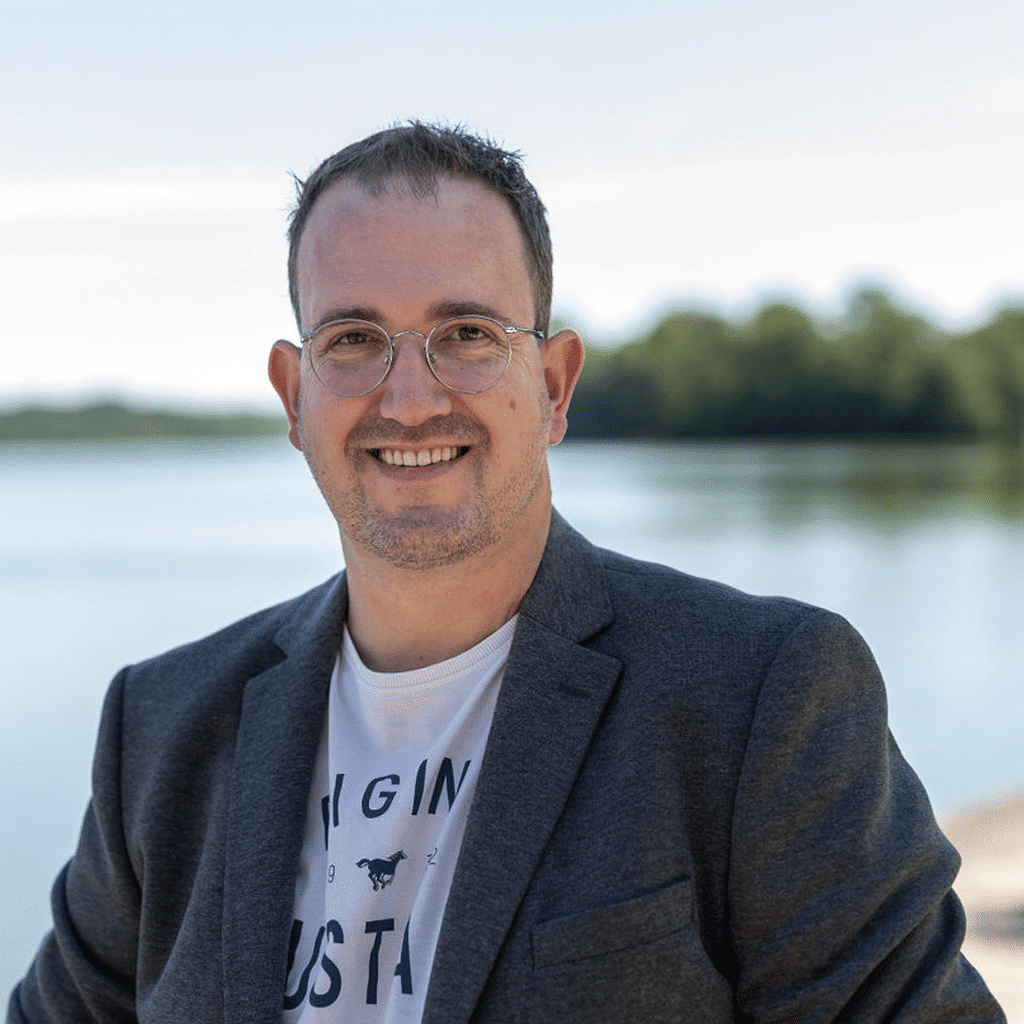 Portraitfoto von Geschäftsinhaber Thorben Junge. Er trägt ein grauesJacket, ein weißes T-Shirt, hat kurze dunkle Haare, freundlicher Blick und im Hintergrund ein See und Wald mit Tiefenschärfe.